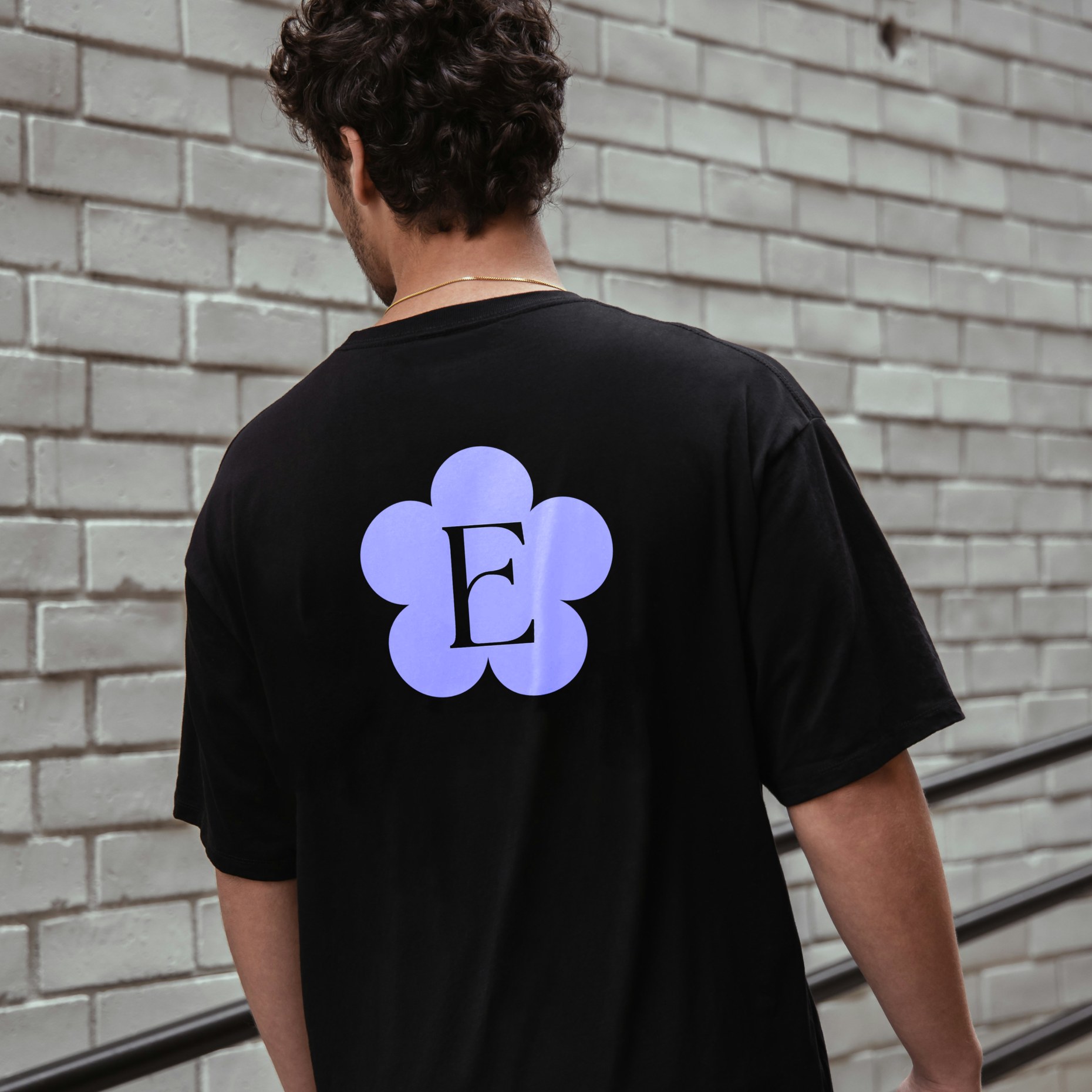 Black t-shirt printed with the lilac Eden flower monogram, worn against a painted brick wall